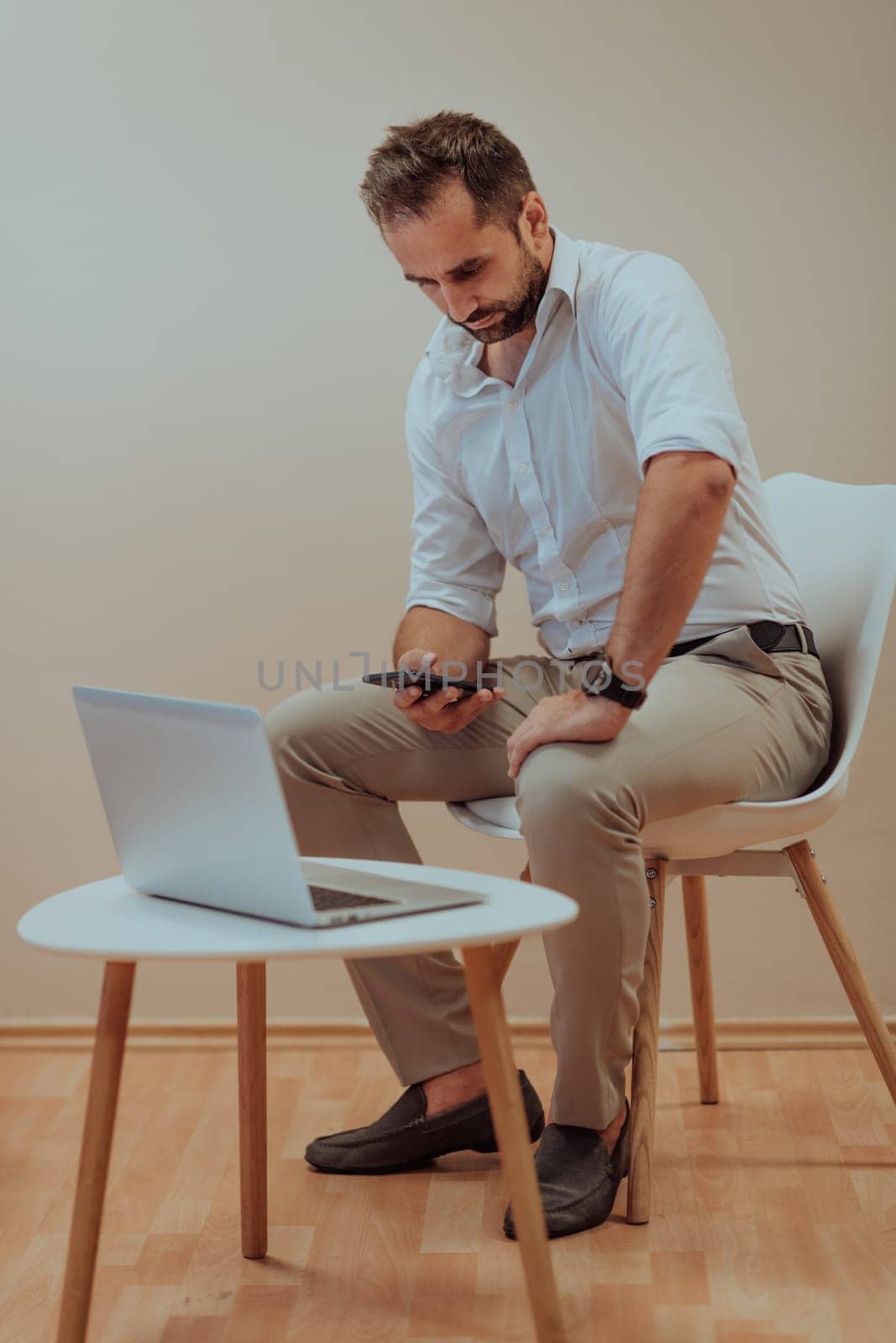 A confident businessman sitting and using laptop and smartphone with a determined expression, while a beige background enhances the professional atmosphere, showcasing his productivity and expertise. by dotshock