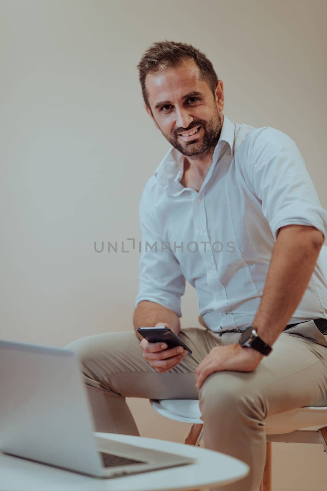 A confident businessman sitting and using laptop and smartphone with a determined expression, while a beige background enhances the professional atmosphere, showcasing his productivity and expertise