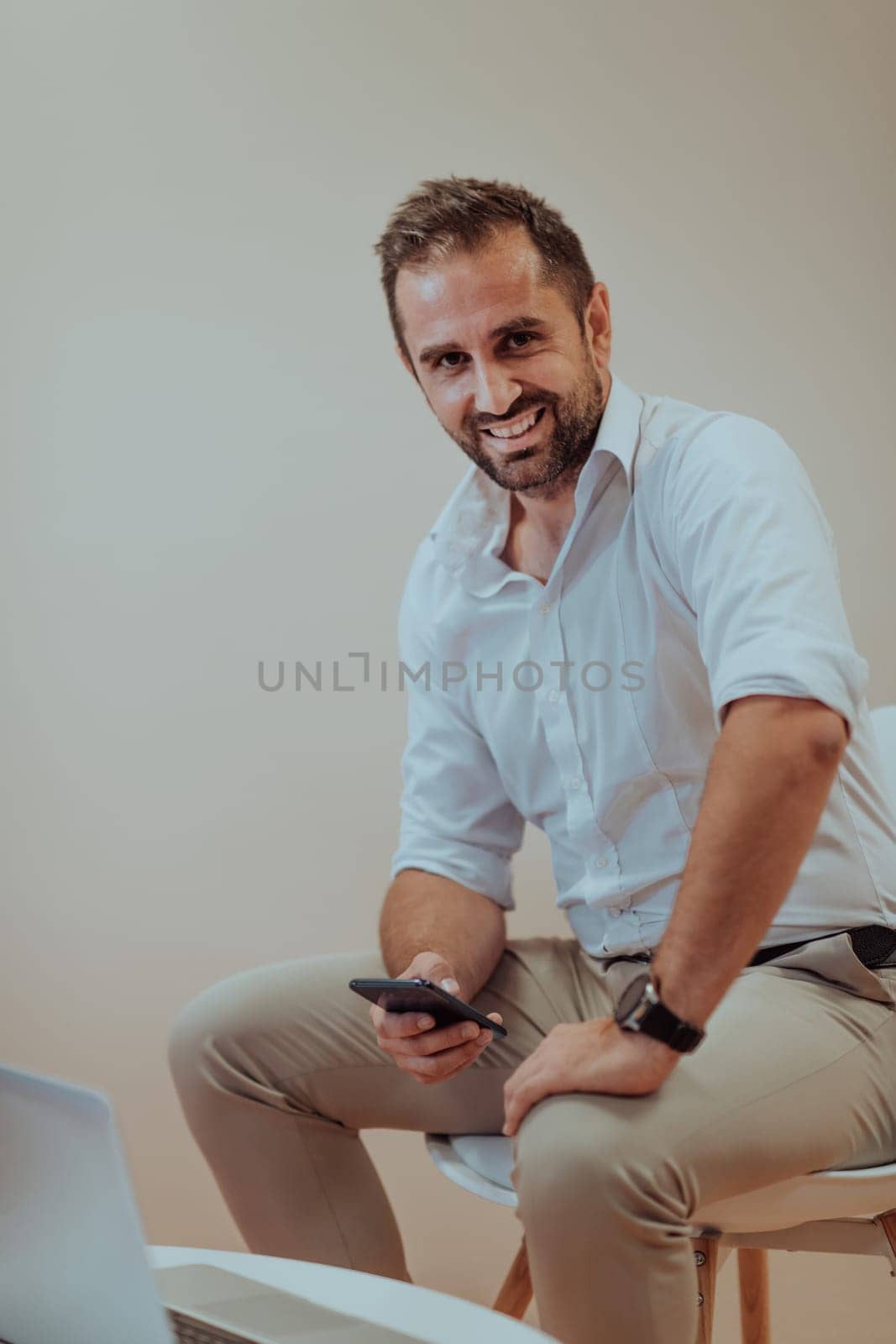 A confident businessman sitting and using laptop and smartphone with a determined expression, while a beige background enhances the professional atmosphere, showcasing his productivity and expertise