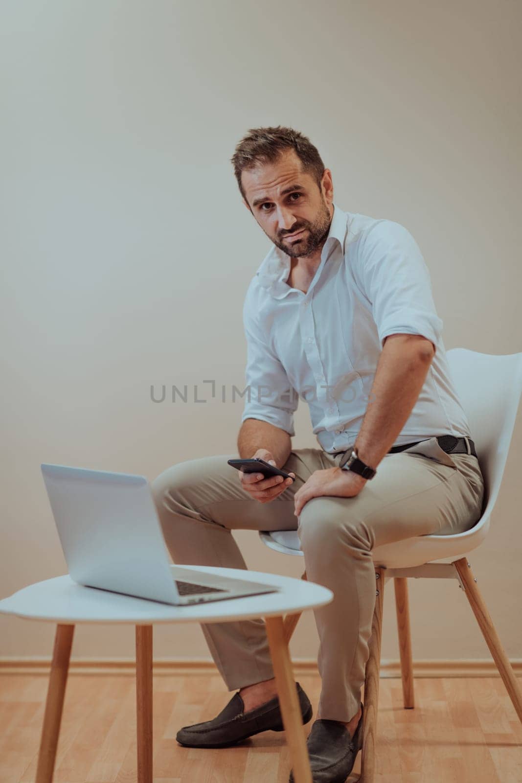 A confident businessman sitting and using laptop and smartphone with a determined expression, while a beige background enhances the professional atmosphere, showcasing his productivity and expertise