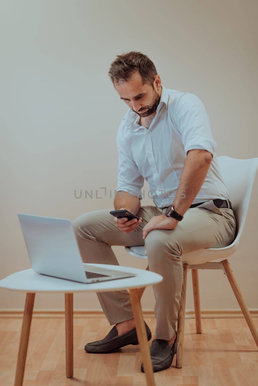 A confident businessman sitting and using laptop and smartphone with a determined expression, while a beige background enhances the professional atmosphere, showcasing his productivity and expertise
