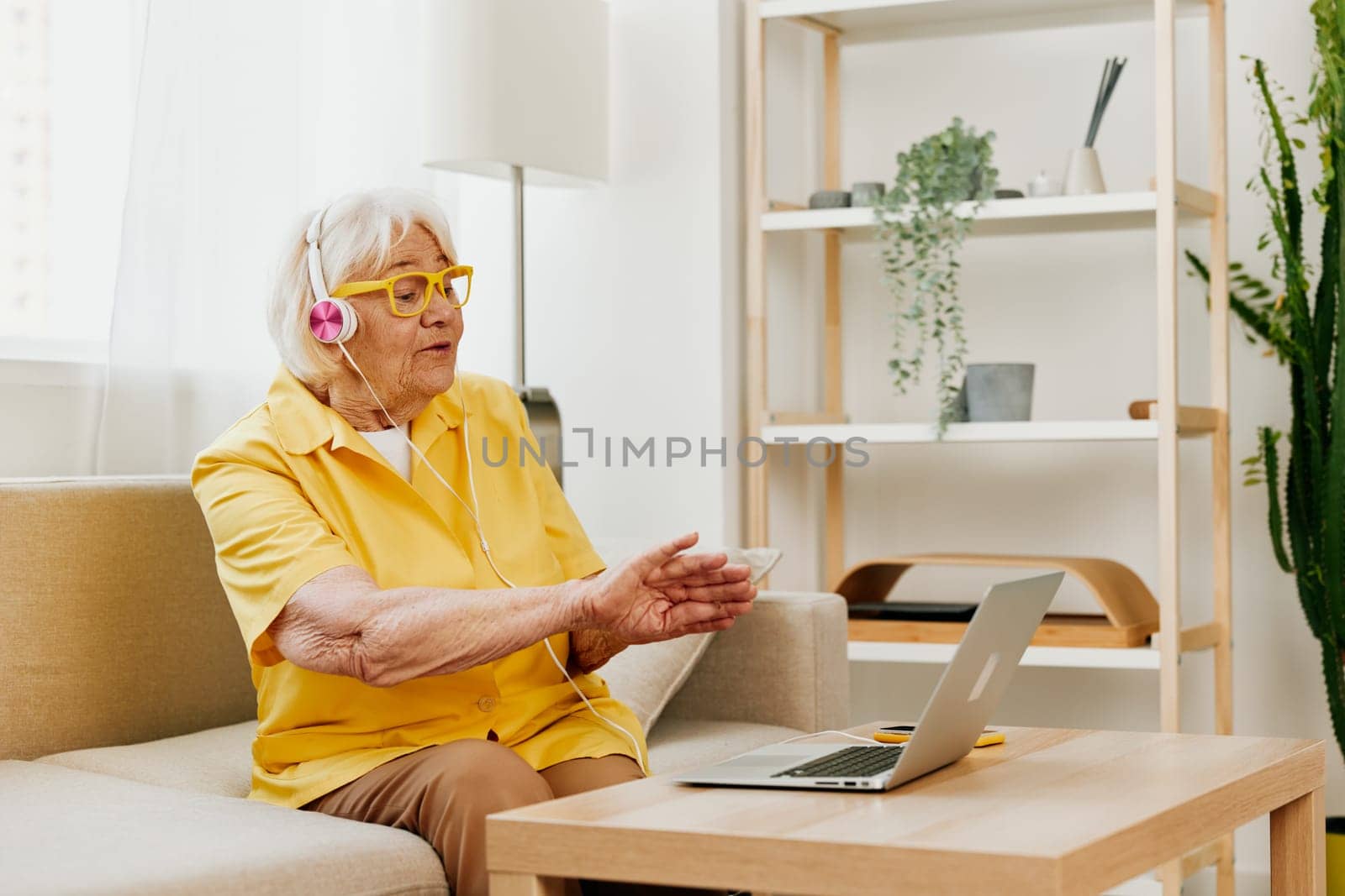 Elderly woman wearing headphones with a laptop communication online video call smile happiness, sitting on the couch at home and working in a yellow shirt, the lifestyle of a retired woman. High quality photo