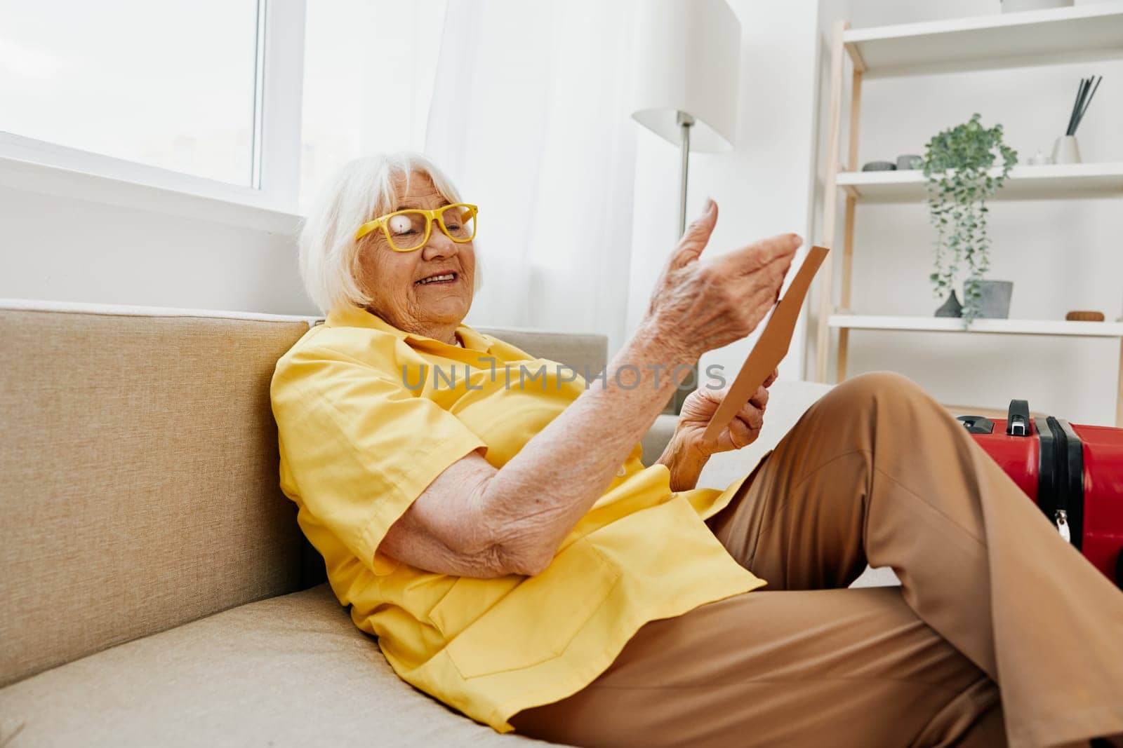 Happy senior woman with passport and travel ticket packed a red suitcase, vacation and health care. Smiling old woman joyfully sitting on the sofa before the trip raised her hands up in joy. High quality photo