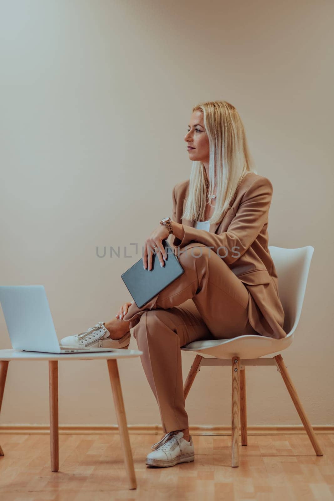 A professional businesswoman sits on a chair, surrounded by a serene beige background, diligently working on her laptop, showcasing dedication and focus in her pursuit of success.