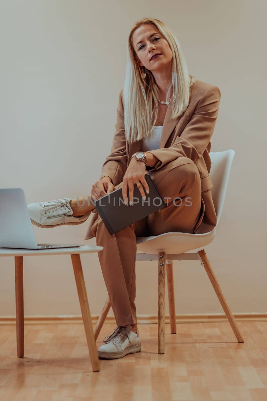 A professional businesswoman sits on a chair, surrounded by a serene beige background, diligently working on her laptop, showcasing dedication and focus in her pursuit of success.