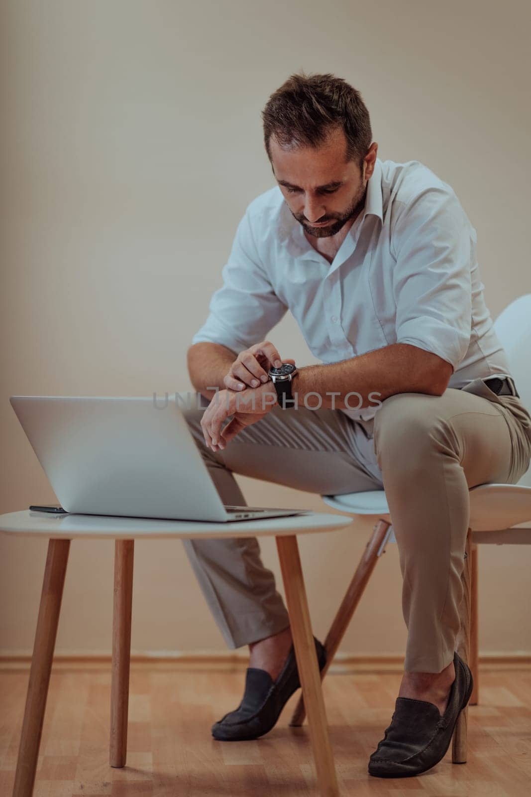 A confident businessman sitting and using laptop and smartwatch with a determined expression, while a beige background enhances the professional atmosphere, showcasing his productivity and expertise