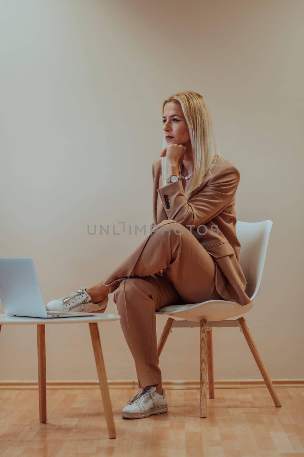 A professional businesswoman sits on a chair, surrounded by a serene beige background, diligently working on her laptop, showcasing dedication and focus in her pursuit of success by dotshock