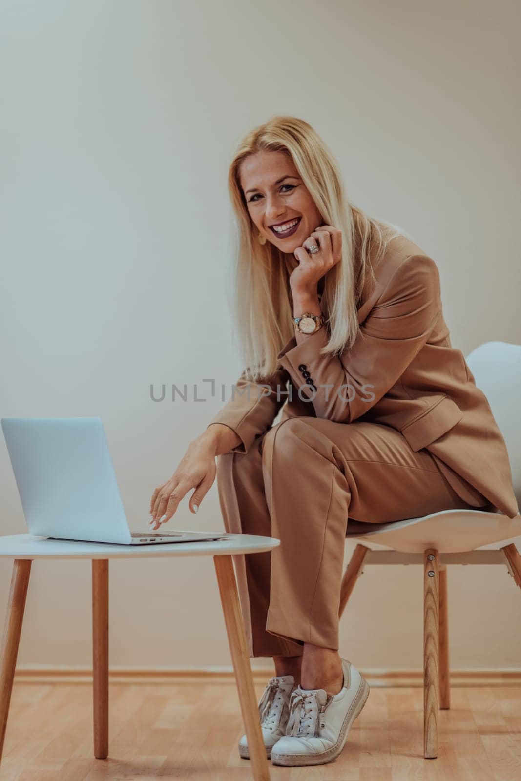 A professional businesswoman sits on a chair, surrounded by a serene beige background, diligently working on her laptop, showcasing dedication and focus in her pursuit of success.