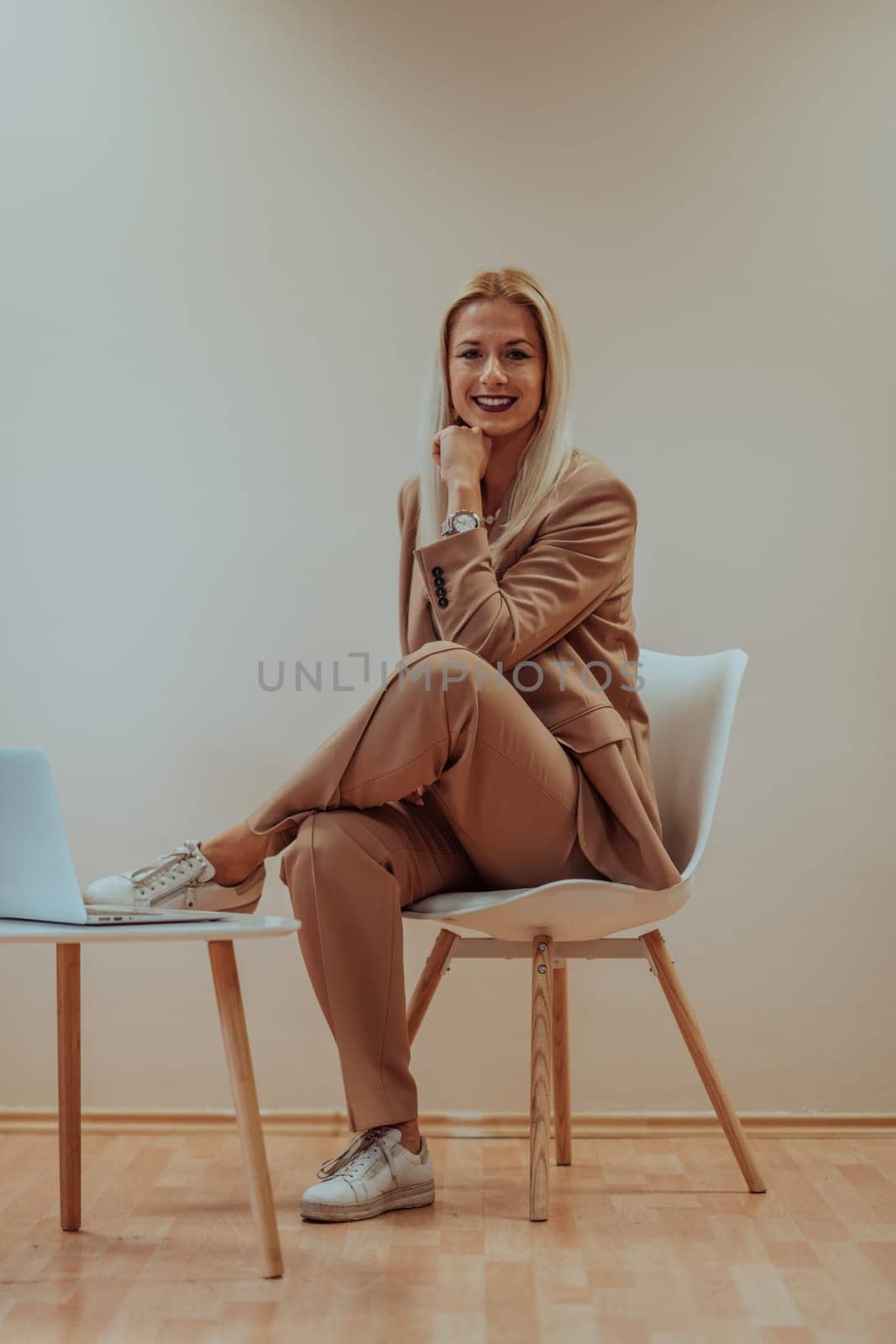 A professional businesswoman sits on a chair, surrounded by a serene beige background, diligently working on her laptop, showcasing dedication and focus in her pursuit of success.