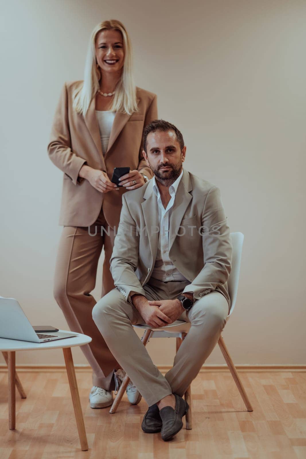 A business couple poses for a photograph together against a beige backdrop, capturing their professional partnership and creating a timeless image of unity and success