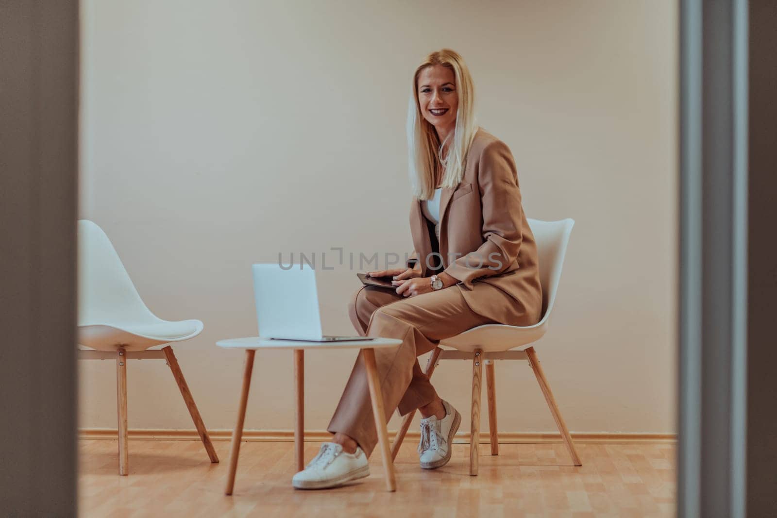 A professional businesswoman sits on a chair, surrounded by a serene beige background, diligently working on her laptop, showcasing dedication and focus in her pursuit of success.