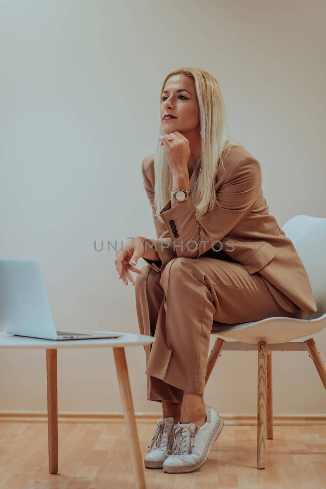 A professional businesswoman sits on a chair, surrounded by a serene beige background, diligently working on her laptop, showcasing dedication and focus in her pursuit of success.