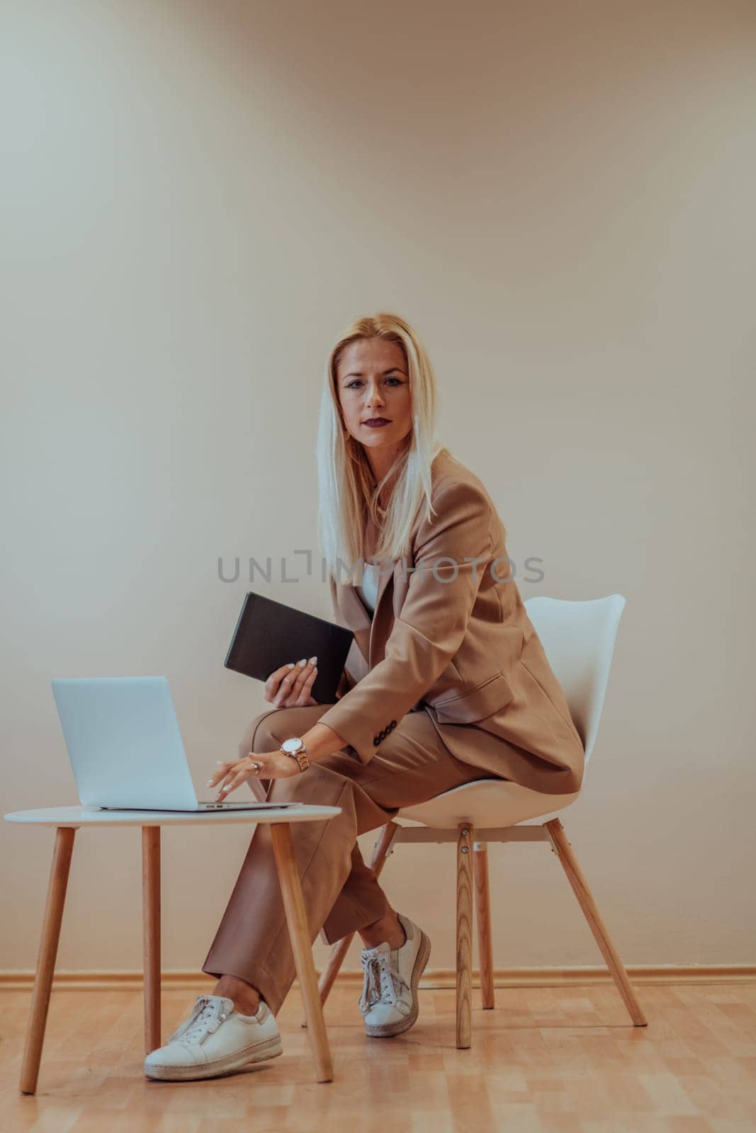 A professional businesswoman sits on a chair, surrounded by a serene beige background, diligently working on her laptop, showcasing dedication and focus in her pursuit of success.