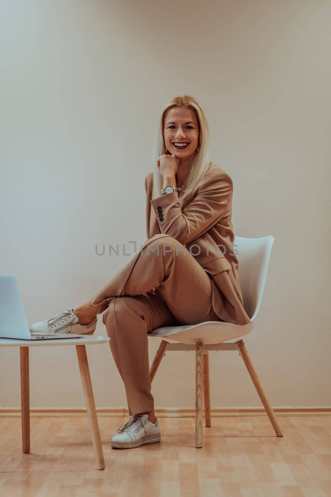 A professional businesswoman sits on a chair, surrounded by a serene beige background, diligently working on her laptop, showcasing dedication and focus in her pursuit of success.