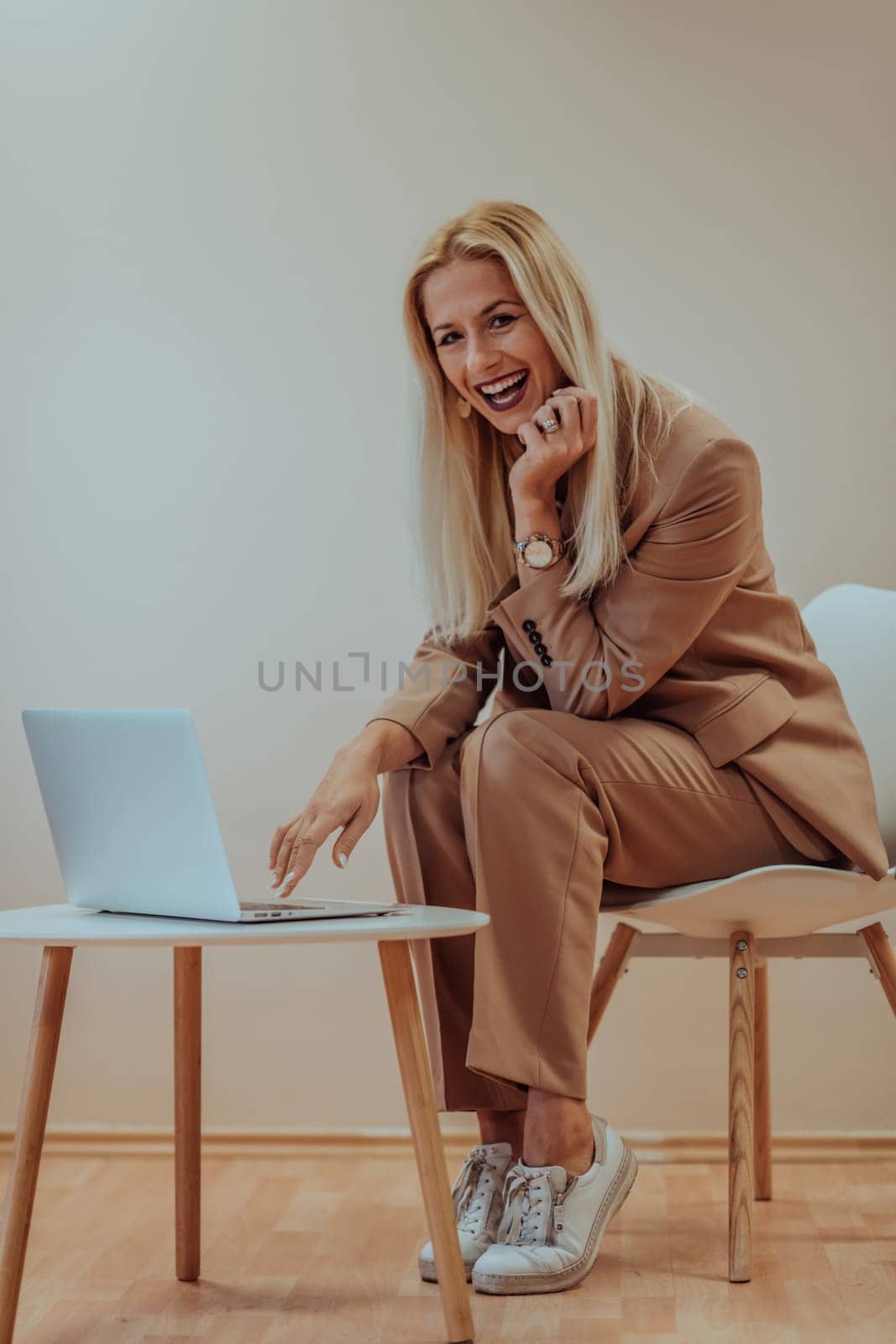 A professional businesswoman sits on a chair, surrounded by a serene beige background, diligently working on her laptop, showcasing dedication and focus in her pursuit of success by dotshock