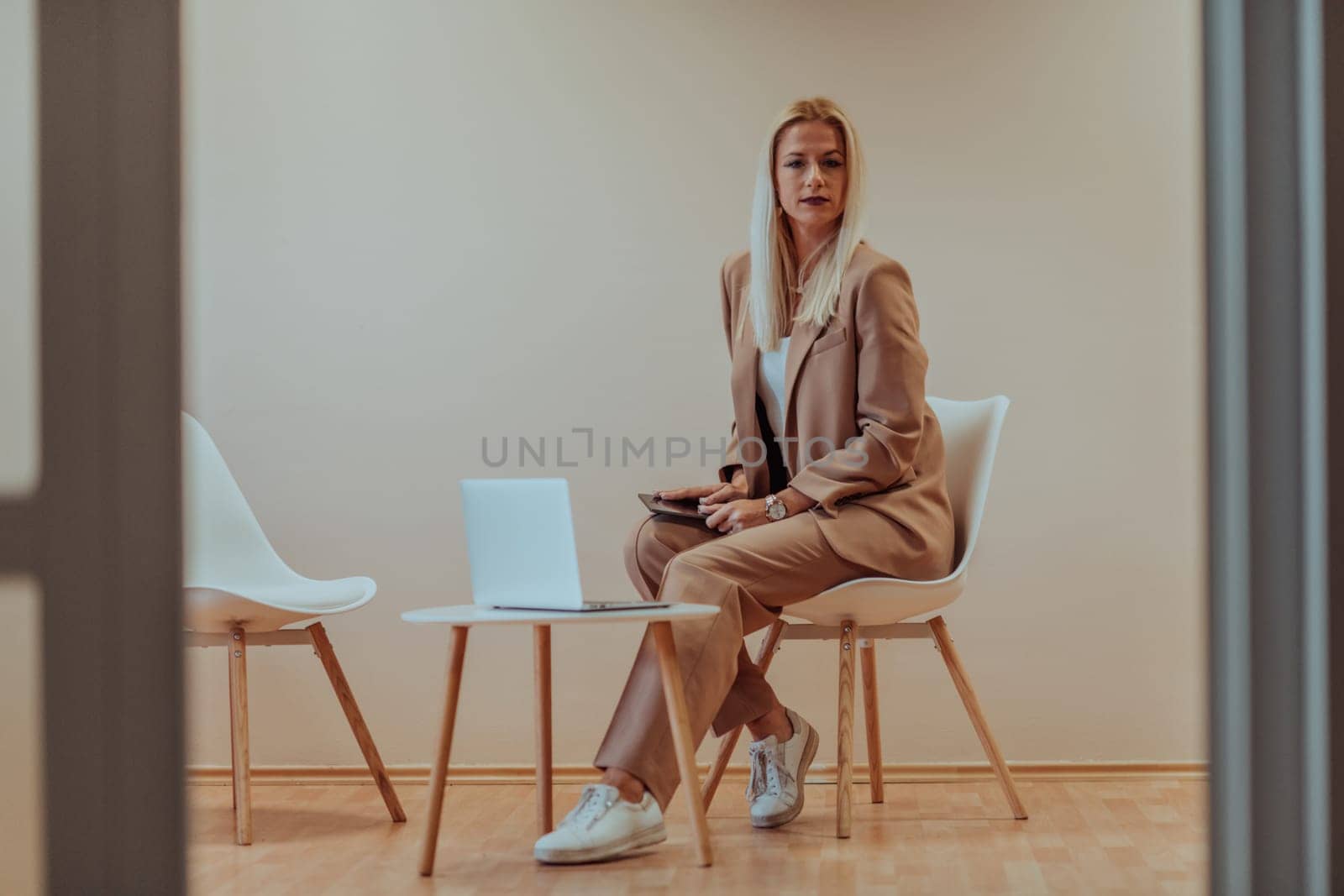 A professional businesswoman sits on a chair, surrounded by a serene beige background, diligently working on her laptop, showcasing dedication and focus in her pursuit of success.