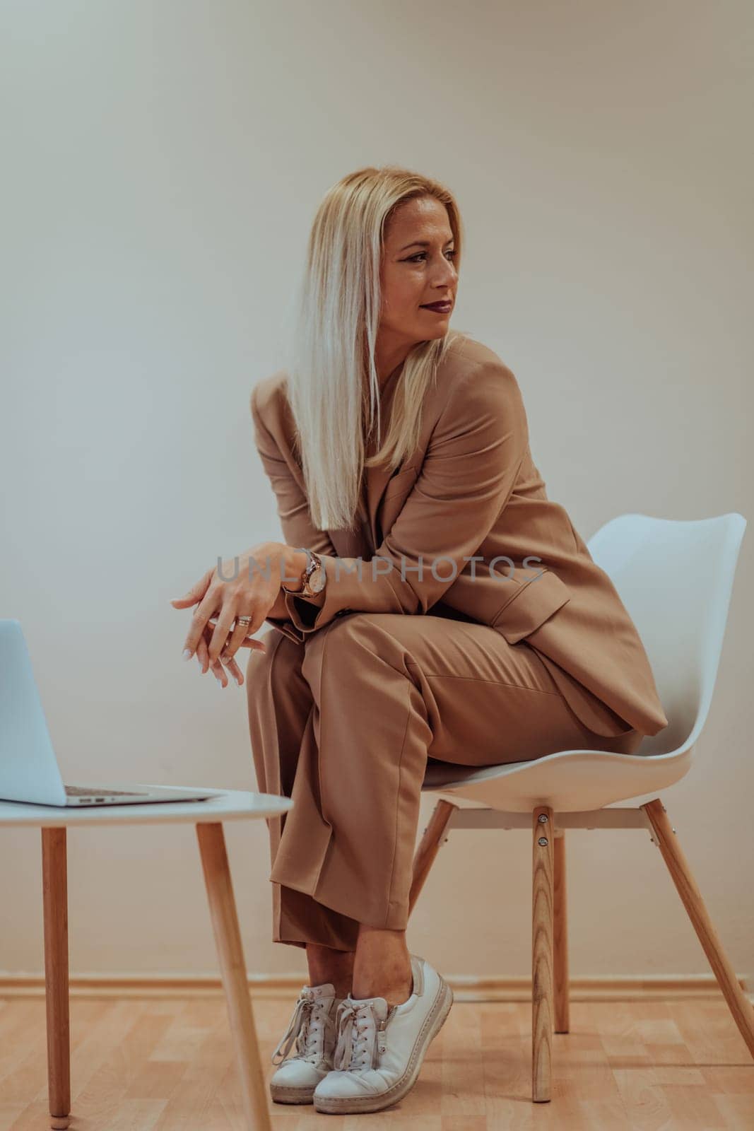 A professional businesswoman sits on a chair, surrounded by a serene beige background, diligently working on her laptop, showcasing dedication and focus in her pursuit of success.