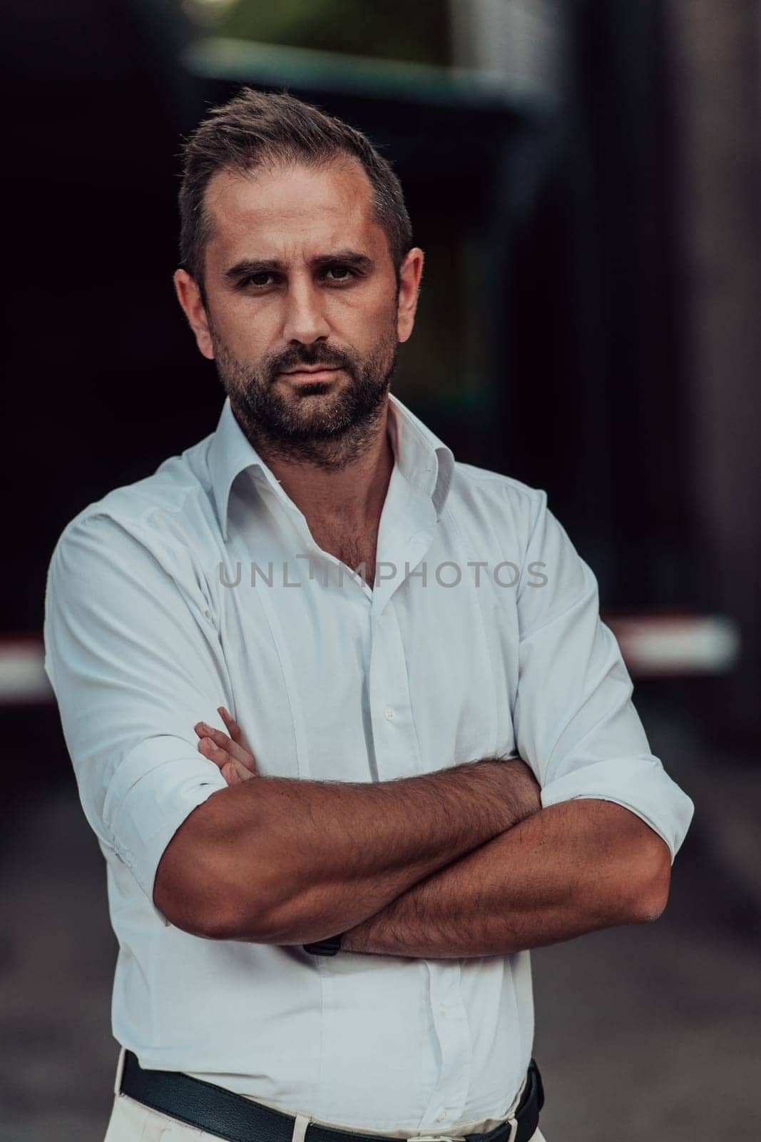 A successful businessman in a white shirt, with crossed arms, poses outdoors, confident expression on his face