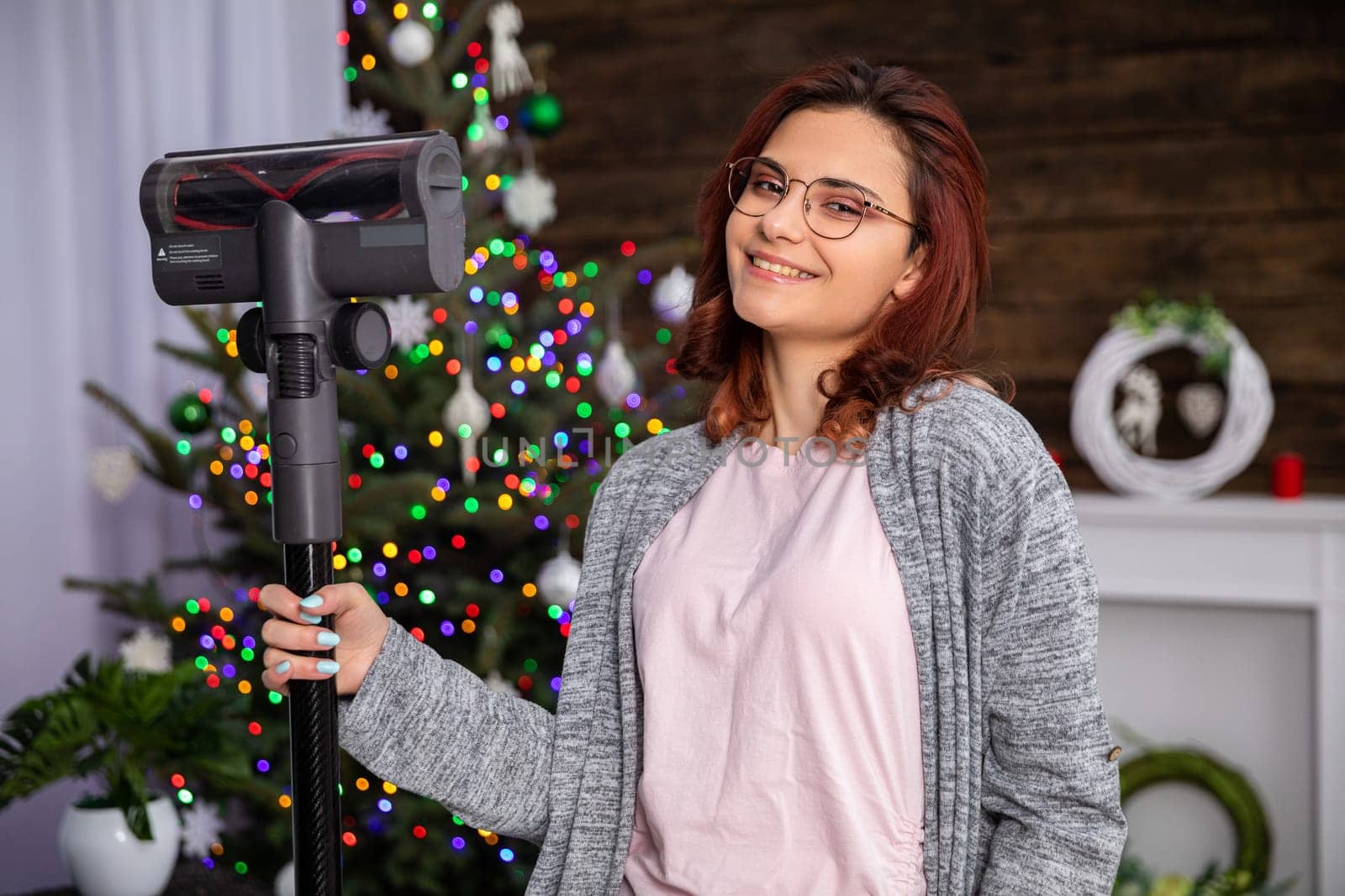 A smiling young woman holds a vacuum cleaner in her hand and a Christmas tree and fireplace can be seen in the background. Domestic cleaning work before Christmas.