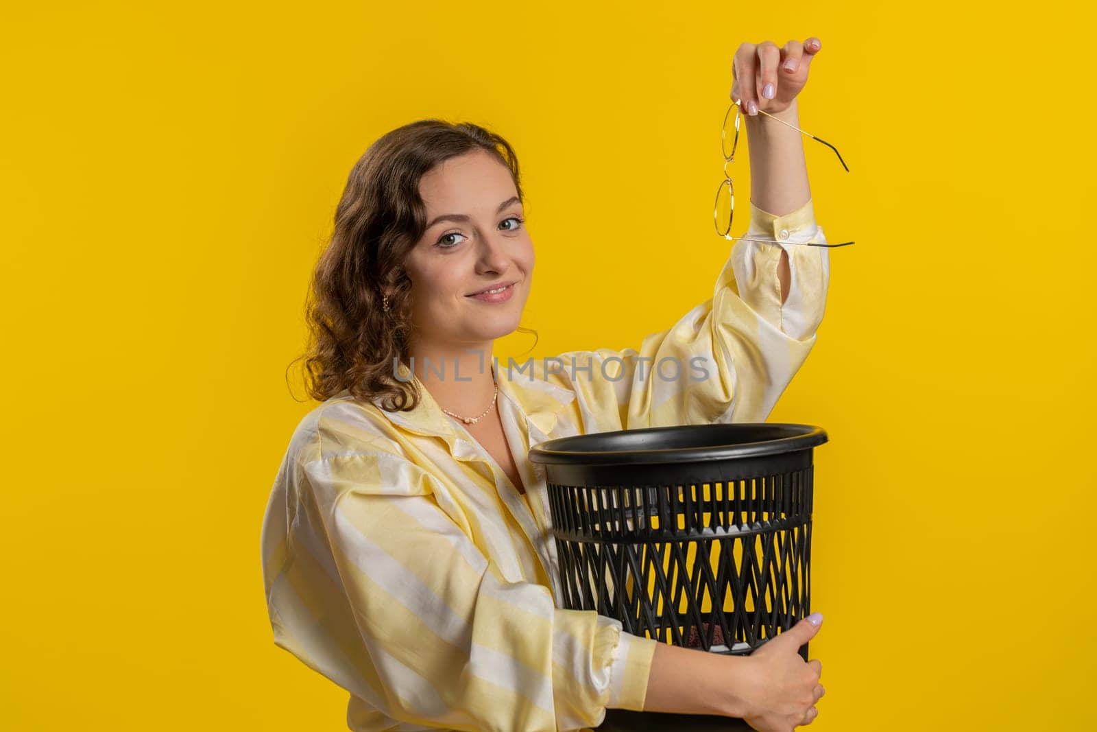 Brunette attractive woman taking off throwing out glasses into bin after medical vision laser treatment therapy surgery looking smiling at camera. Pretty girl isolated on yellow studio background