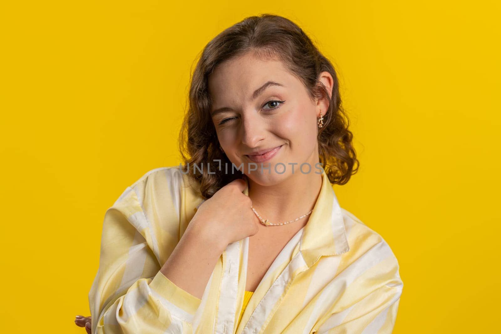 Smiling playful happy young woman blinking eye, looking at camera with smile, winking and flirting, expressing optimism. Excited girl isolated alone on yellow studio background. Lifestyles