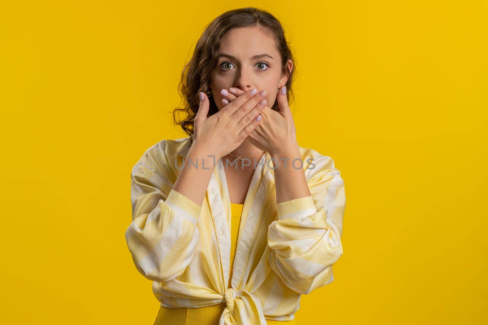 I will not say anything. Frightened woman closing mouth with hands, looking intimidated scared at camera, gestures no, refusing to tell terrible secret, unbelievable truth. Girl on yellow background