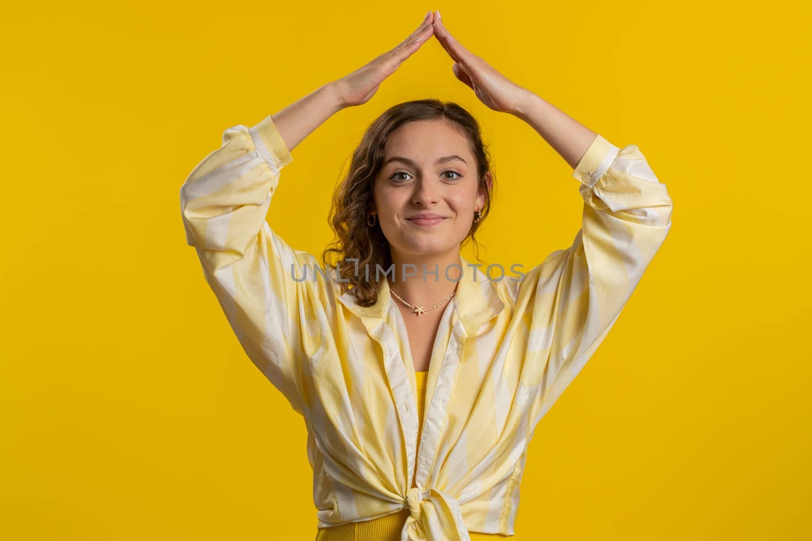 House roof about head. Young woman playing childish catching up game, feeling in safe making roof above head with hands, insurance, security service. Girl isolated alone on yellow studio background
