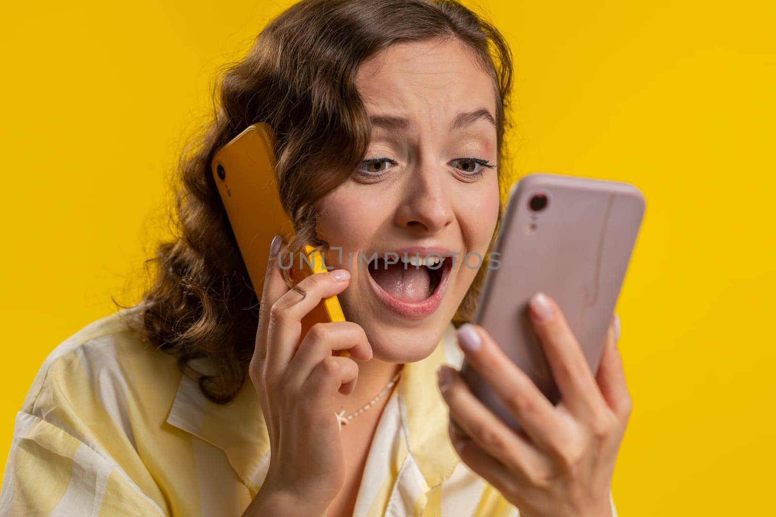 Irritated nervous stressed young woman talking screaming on two mobile phones having conversation conflict quarrel complaint dispute discuss solve problem. Girl isolated on yellow studio background