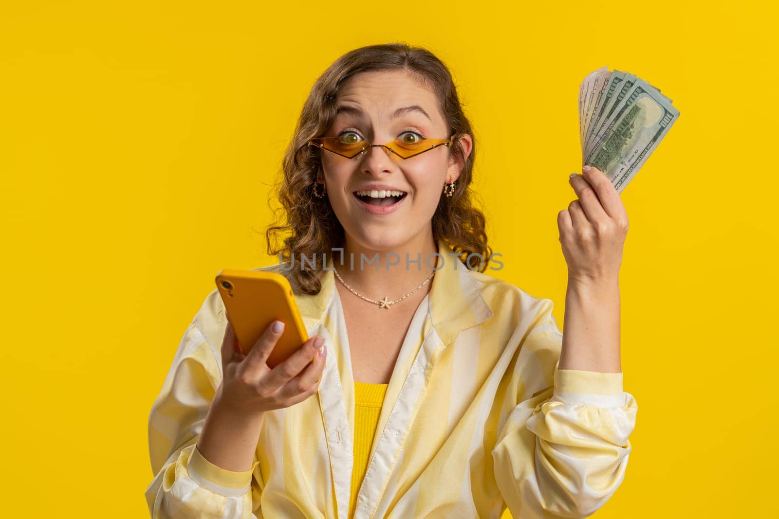 Young woman looking smartphone display sincerely rejoicing win, receiving money dollar cash banknotes, celebrating success lottery luck. Girl isolated alone on yellow studio background. Lifestyles