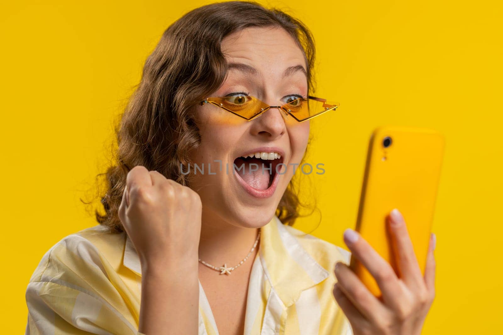 Happy excited joyful woman girl use smartphone typing browsing shouting say wow yes found out great big win, good news, lottery goal achievemen, celebrating success, winning game on yellow background
