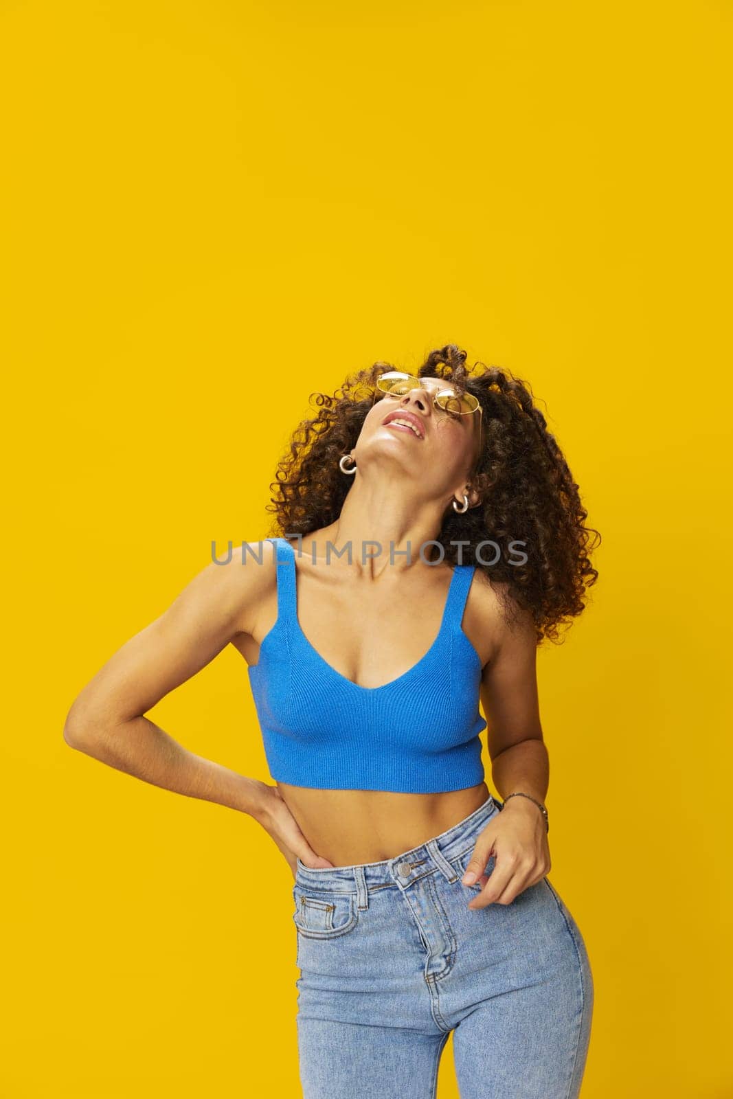 Woman with curly afro hair in a blue T-shirt on a yellow background dancing flying hair with sunglasses yellow, hand signs, look into the camera, smile with teeth and happiness, copy space by SHOTPRIME