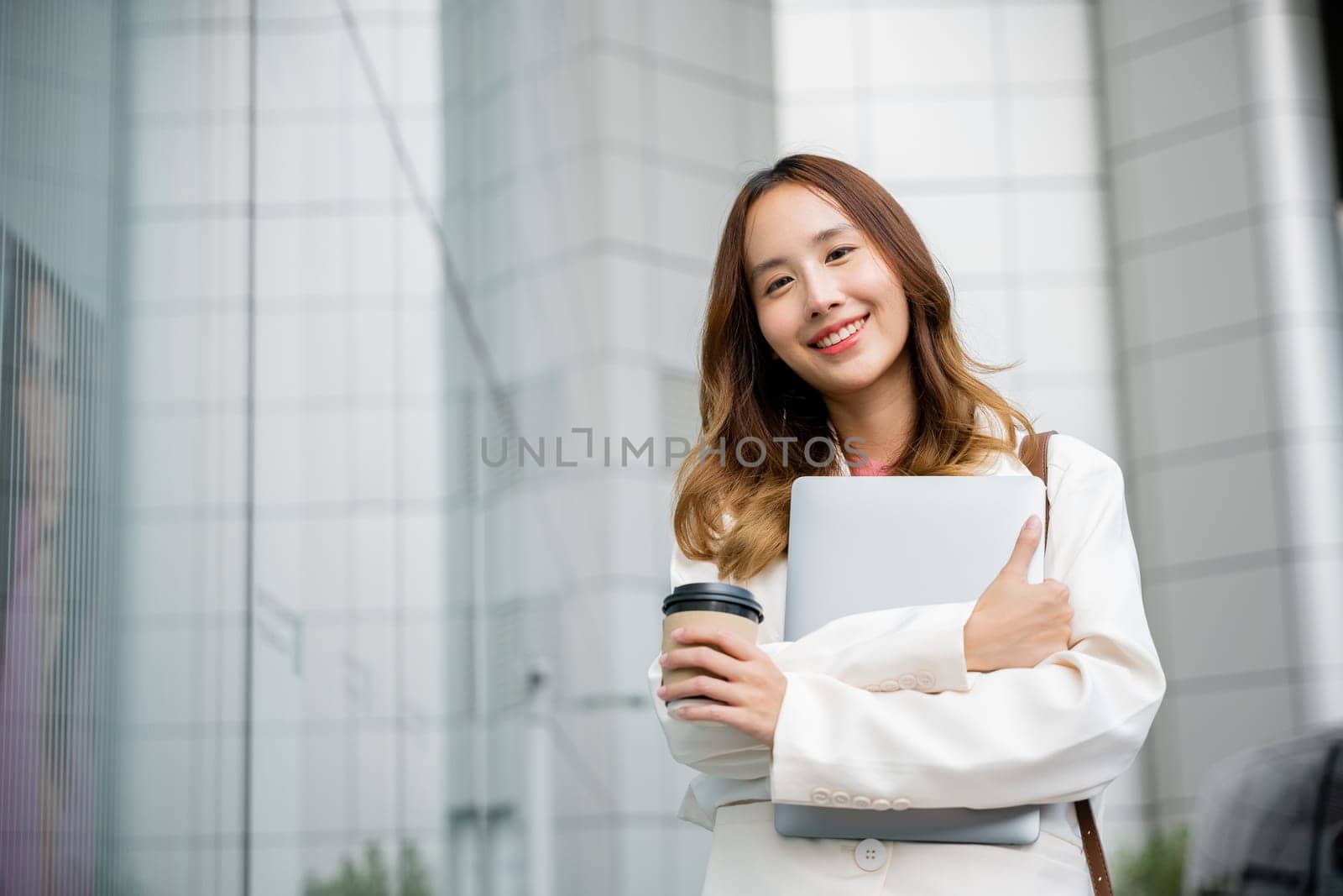 Against the backdrop of a beige wall, a young woman walks through the city with a laptop and a takeaway cup of coffee in her hands. She is smiling, enjoying her digital lifestyle