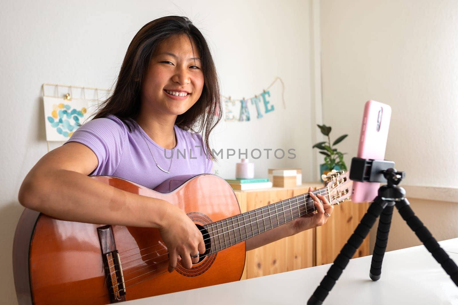 Young Asian woman recording herself playing acoustic guitar with phone. Social media live stream. Asian girl looking at camera. E-learning and hobbies.