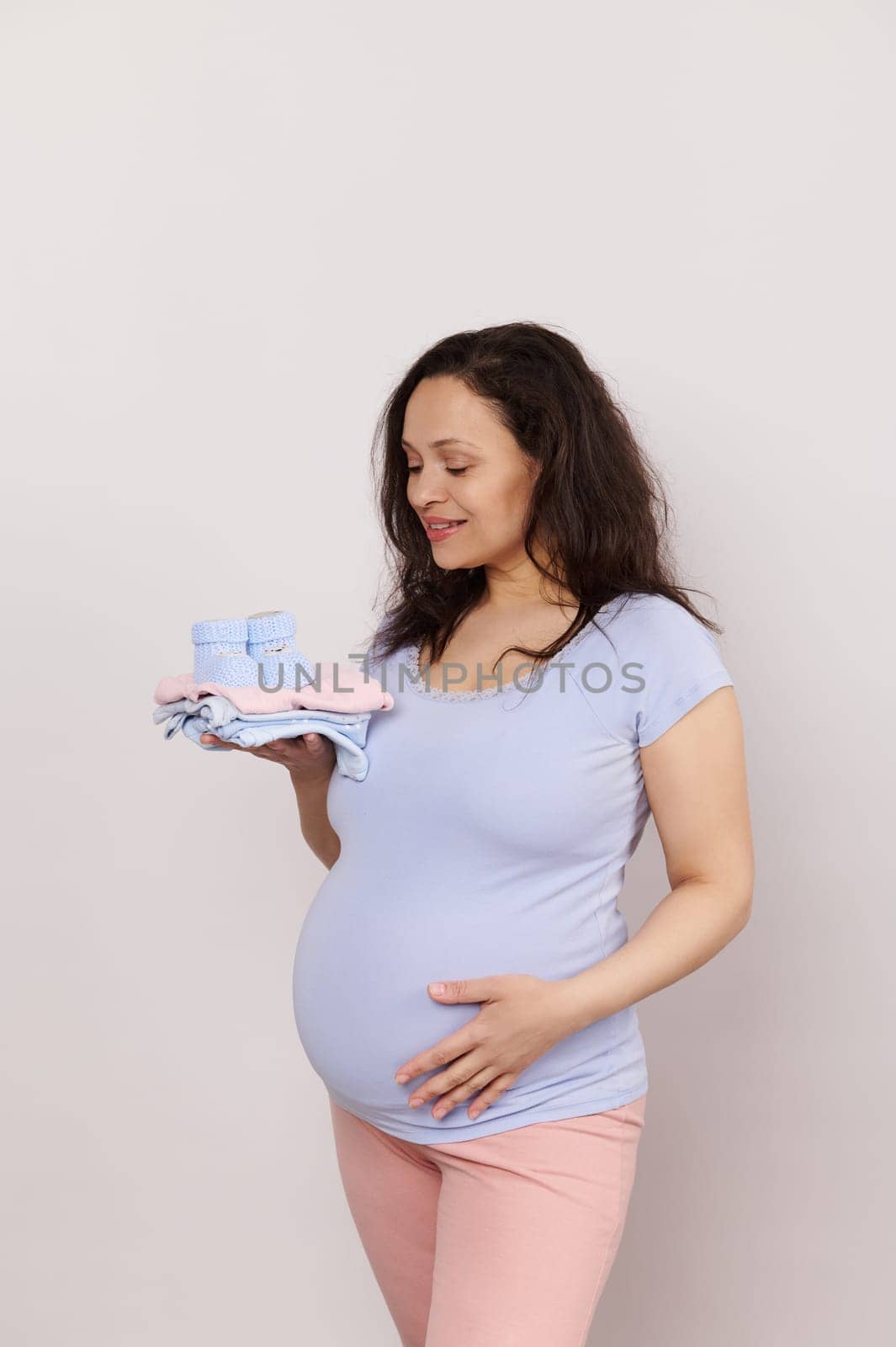 Happy pregnant future mother expecting baby, holds a pile of baby clothes, gently strokes her belly on white isolated background. Gravid female expressing positive emotions of pregnancy and maternity