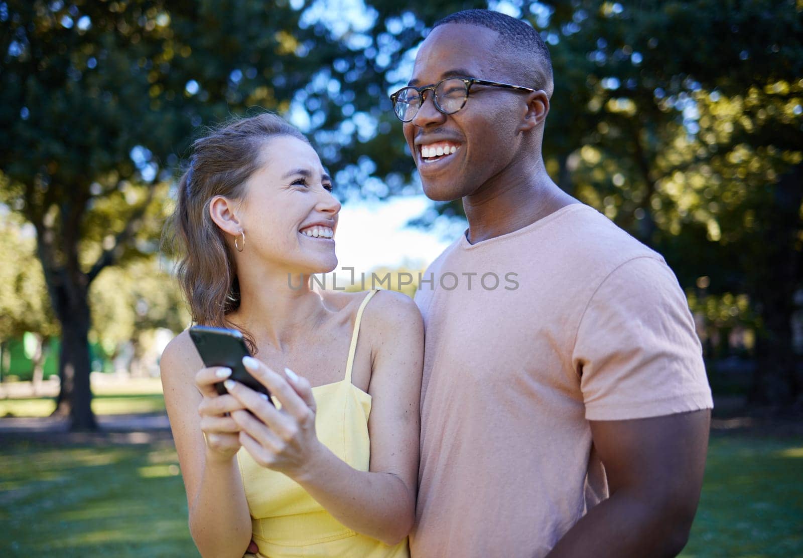 Comedy, laughing and interracial couple with a phone in nature, funny communication and smile at a meme. Comic, streaming and black man and woman reading a joke on a mobile in a park in France by YuriArcurs