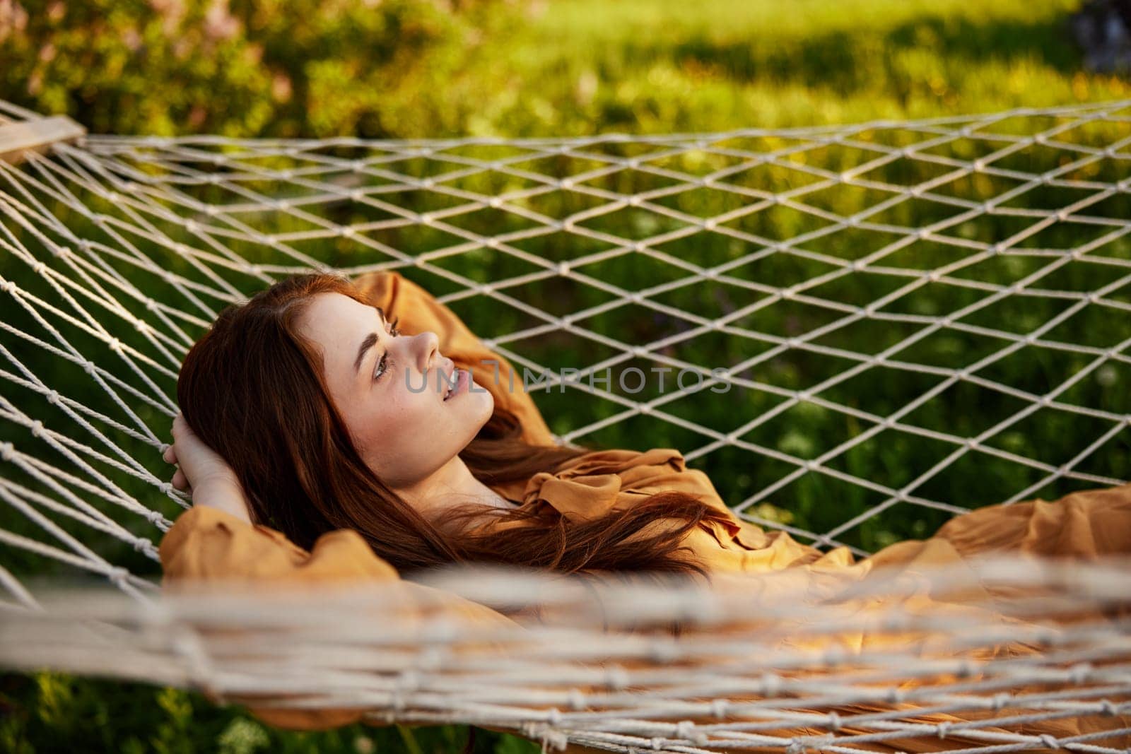 a woman is resting lying in a mesh hammock with her hands behind her head, smiling happily, enjoying a warm day in the rays of the setting sun, lying in an orange dress. High quality photo
