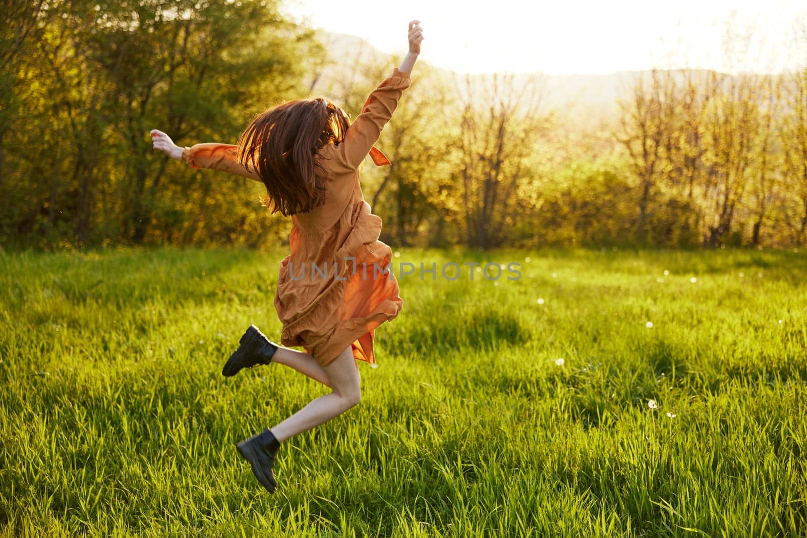 a happy woman in an orange dress illuminated by the rays of the setting sun happily jumps in a green field in the park, enjoying nature and a warm summer day. Horizontal photo. High quality photo