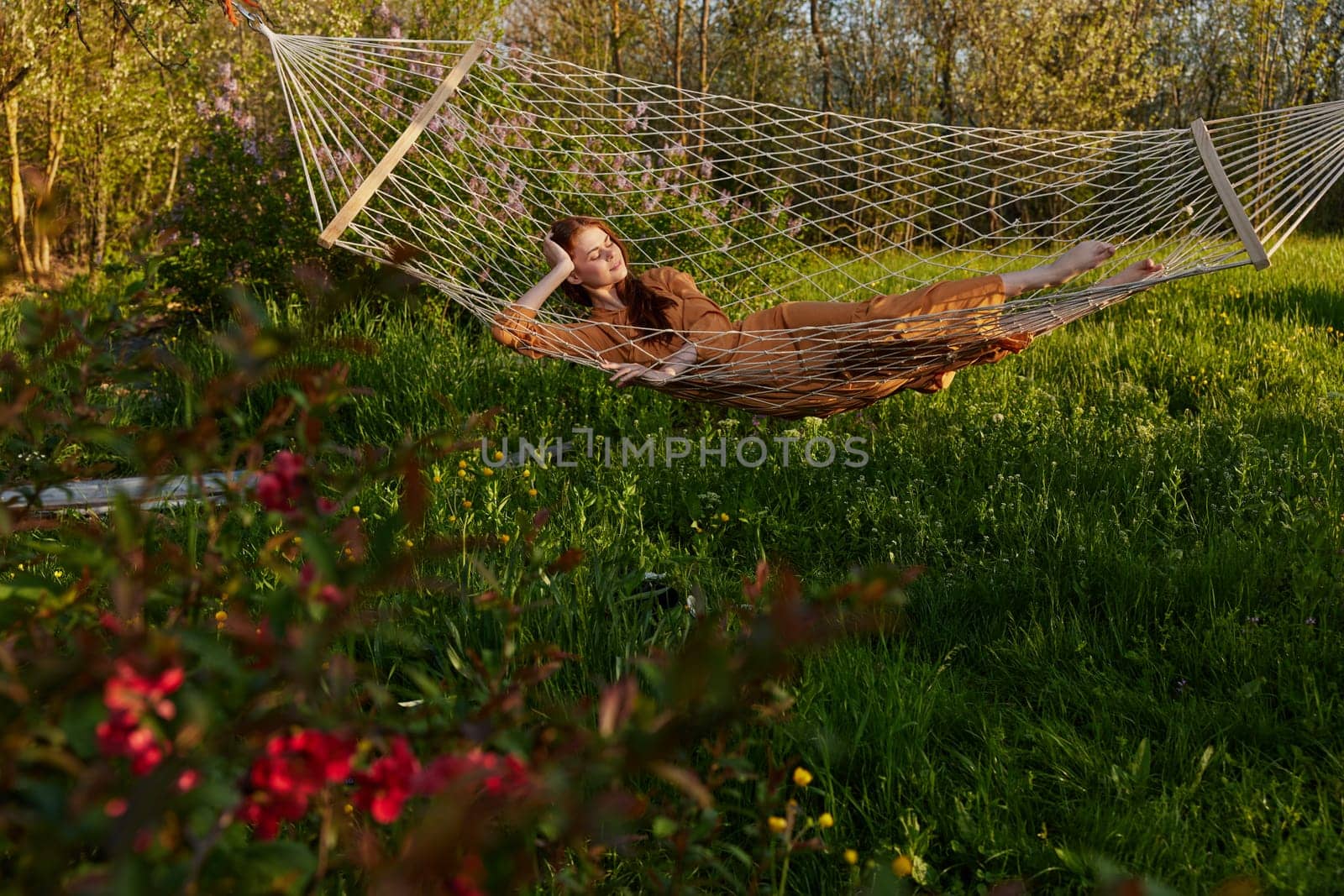 a happy woman in a long orange dress is relaxing in nature lying in a mesh hammock enjoying the peace and tranquility in the country surrounded by green foliage by Vichizh