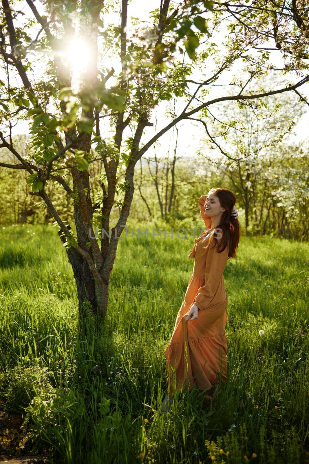 a beautiful, joyful woman stands in a long orange dress, in the countryside, near a tree blooming with white flowers, during sunset, illuminated from behind and touches her long hair by Vichizh