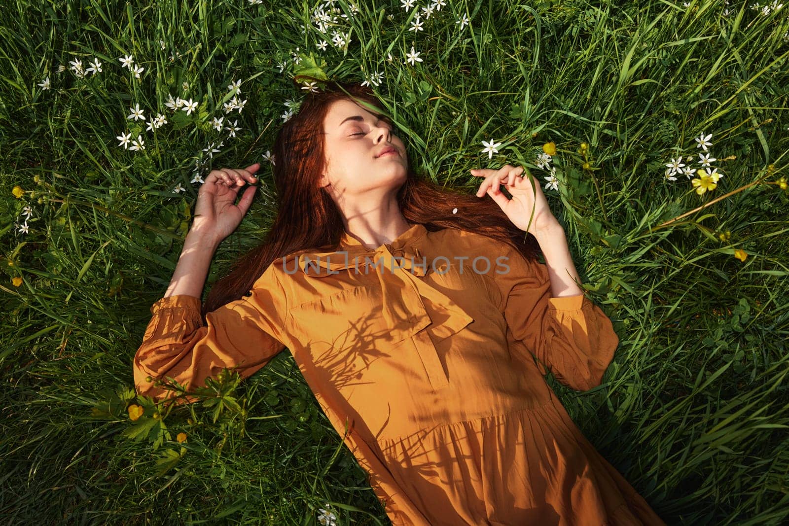 a relaxed woman, resting lying in the green grass, in a long orange dress, with her eyes closed and a pleasant smile on her face, recuperating, illuminated by the warm rays of the setting summer sun. High quality photo