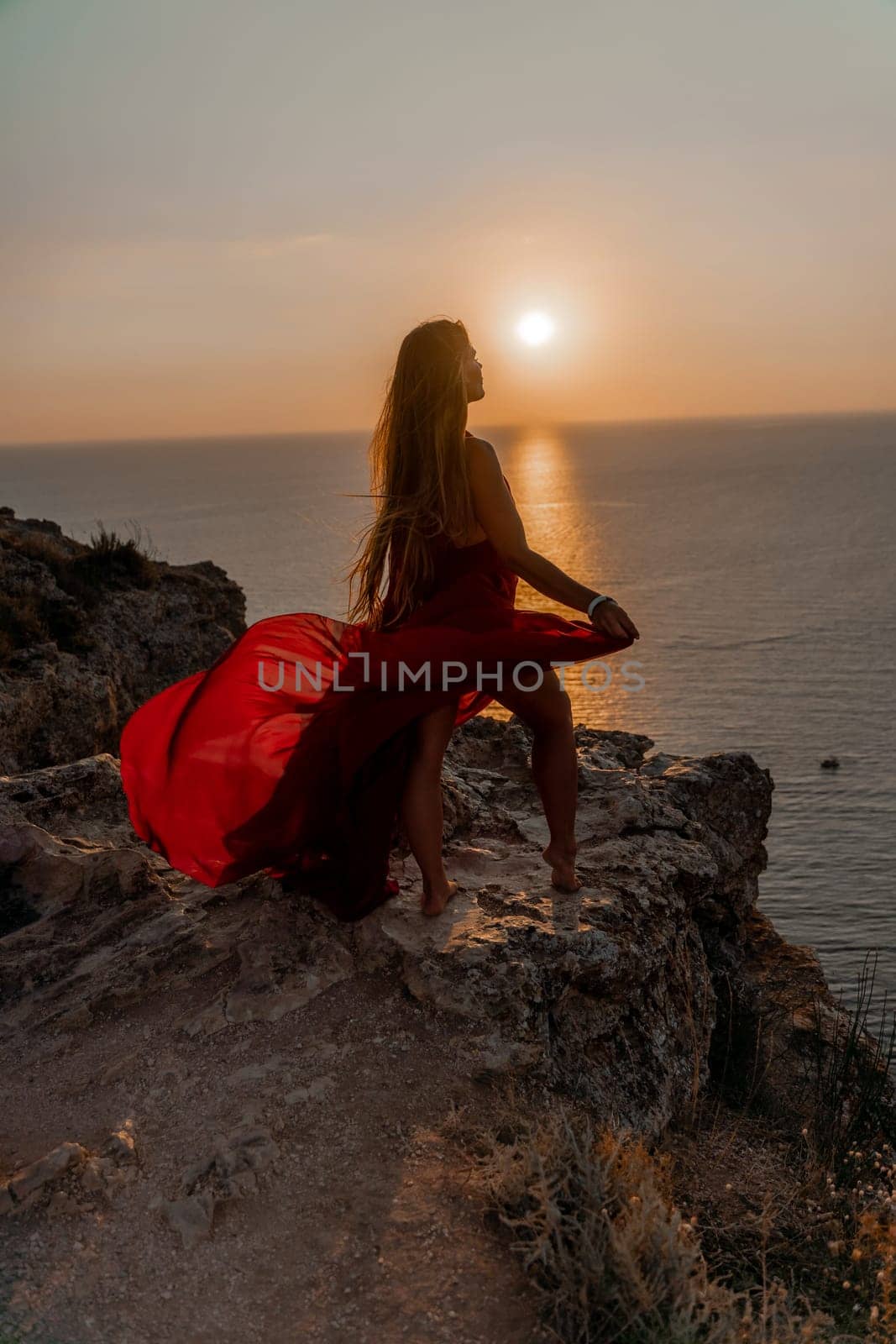 Woman sunset sea red dress, side view a happy beautiful sensual woman in a red long dress posing on a rock high above the sea on sunset