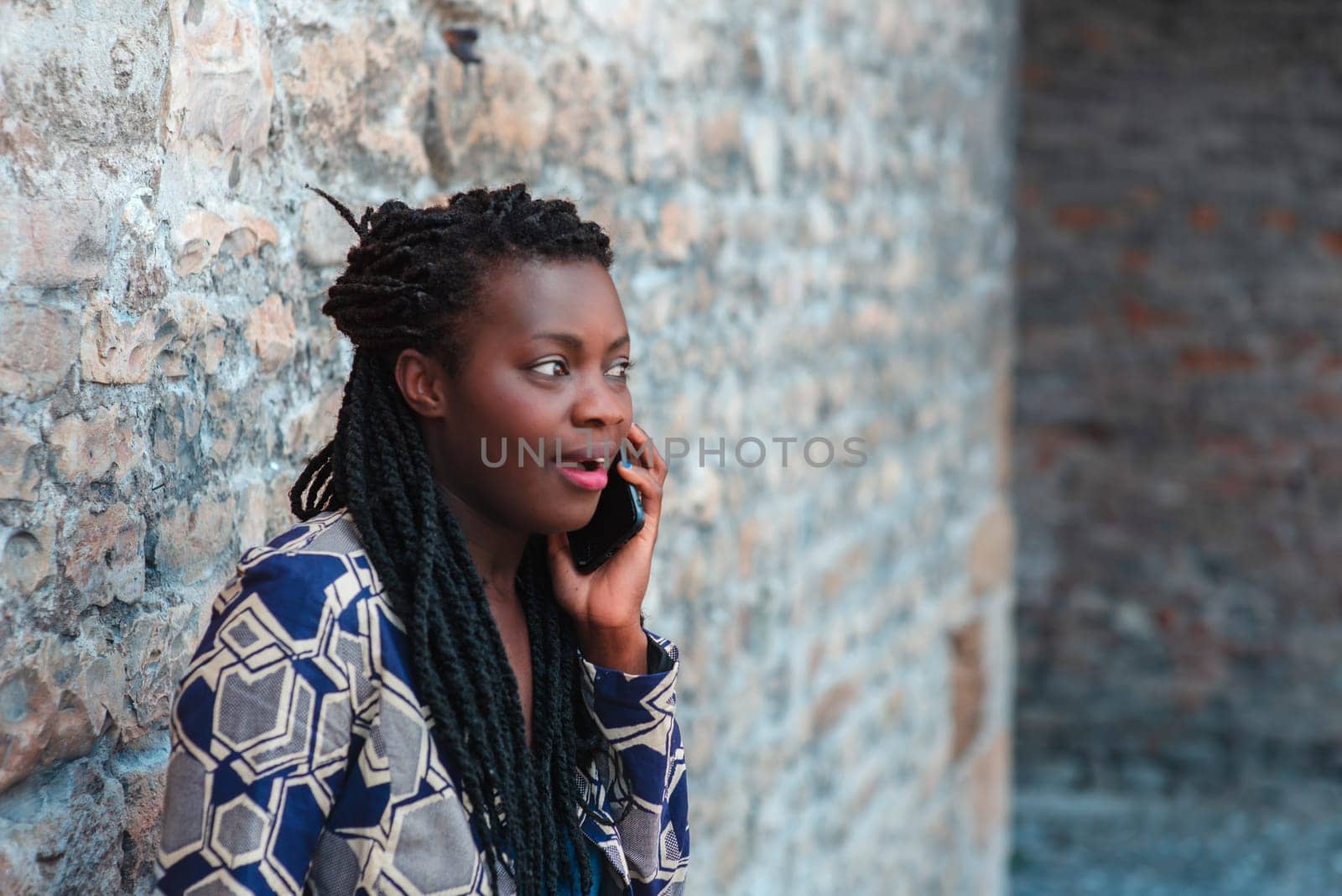 confident African young adult woman holding cell phone calling , receives good news, smiling, happy, outdoors in the village over a wall.