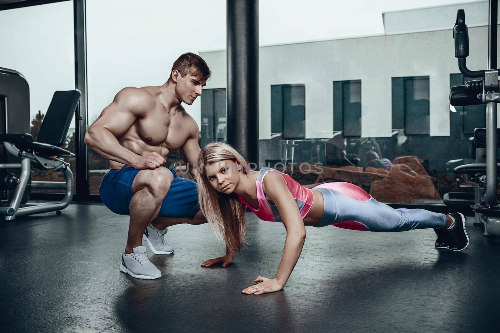 Sporty girl doing plank exercises with support of her personal trainer. Personal trainer with a gorgeous body without a T-shirt in the gym