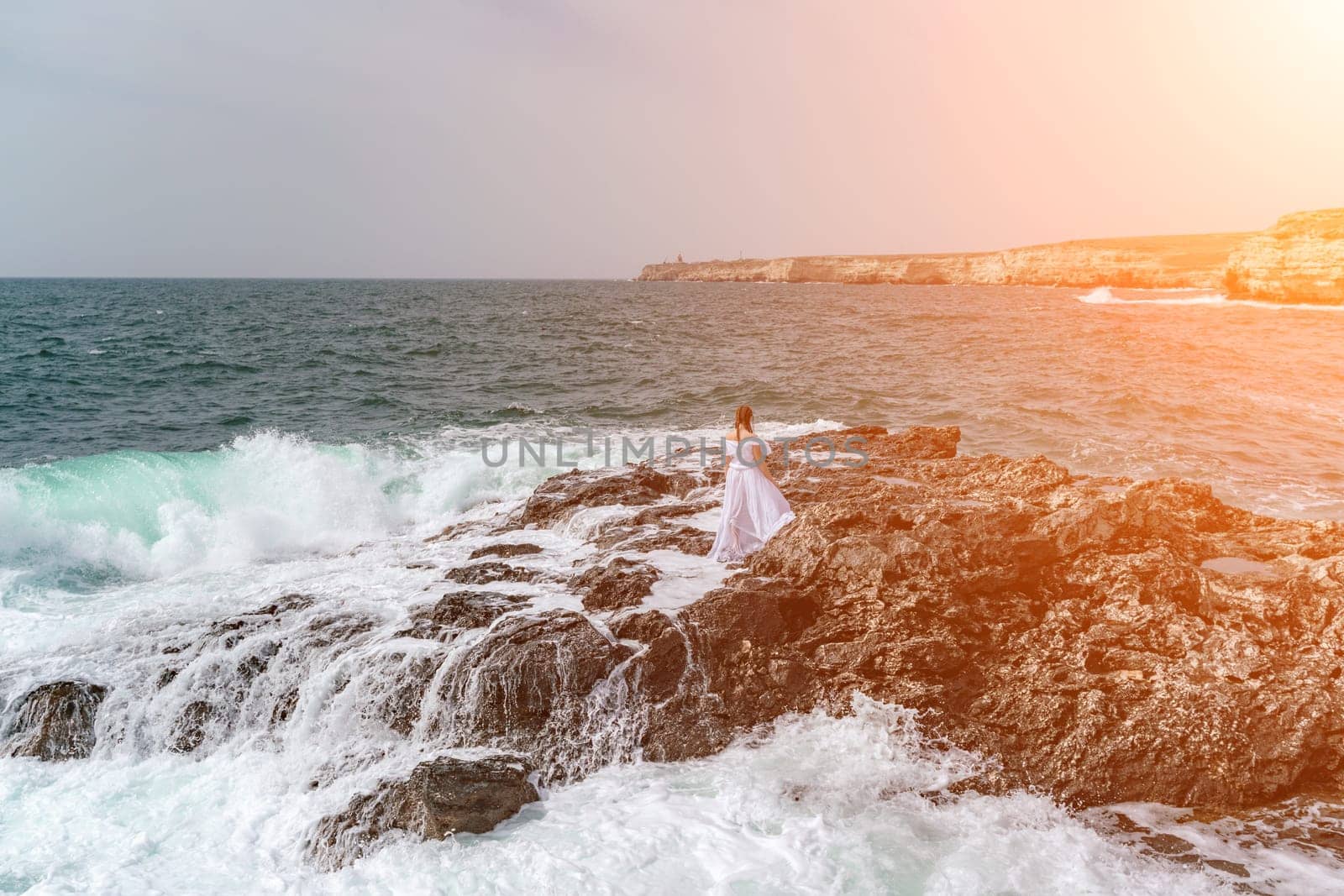 A woman stands on a rock in the sea during a storm. Dressed in a white long dress, the waves break on the rocks and white spray rises