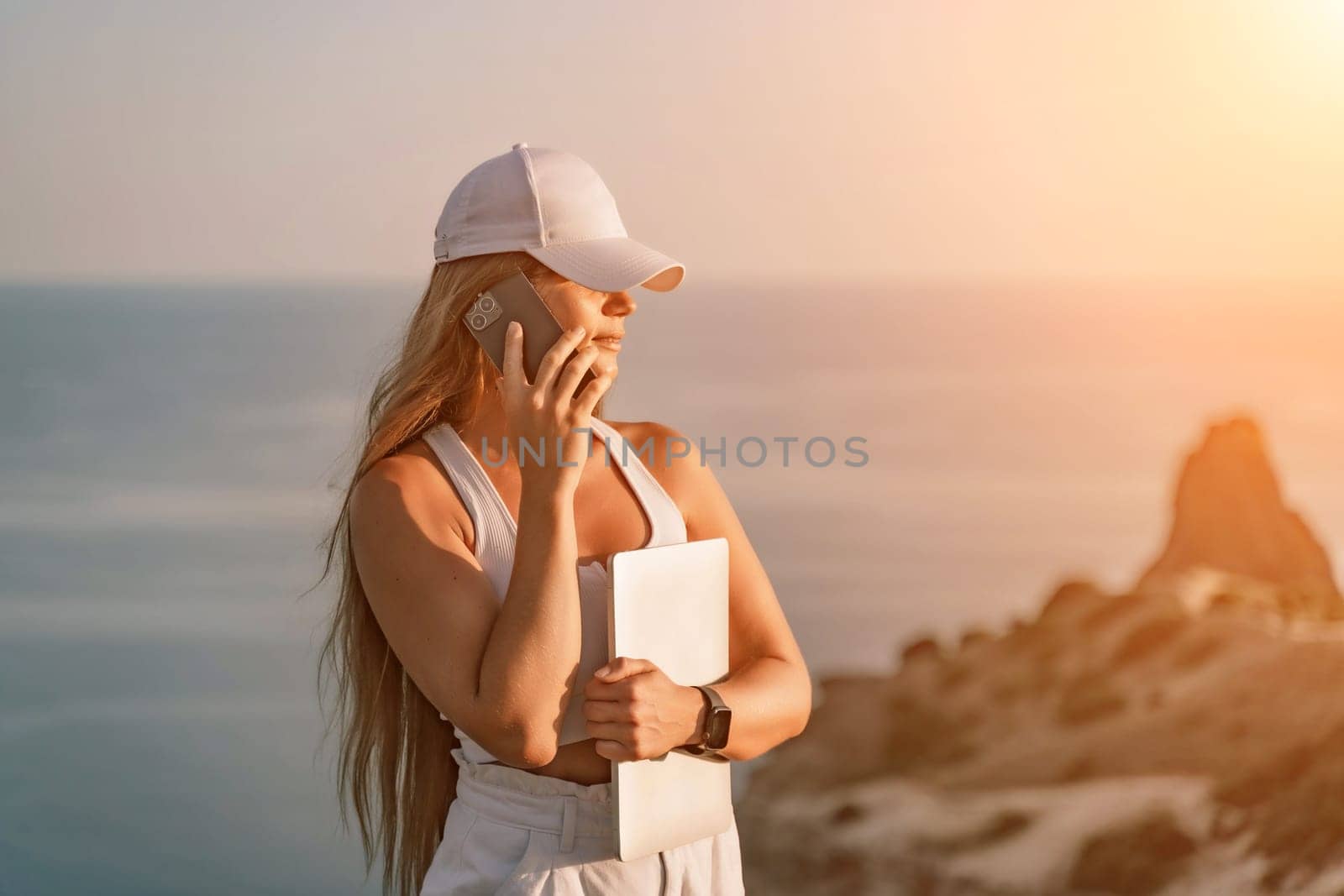 A happy woman in white shorts and T-shirt enjoys the picturesque sea view while holding a laptop and talking on the phone