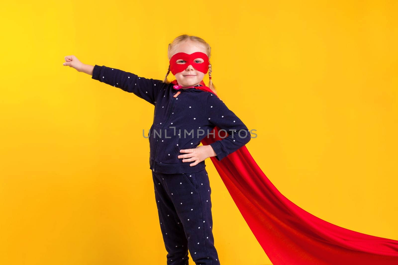 Funny little power superhero child (girl) in a red raincoat and a mask on a yellow background. Studio photography. Superhero concept.