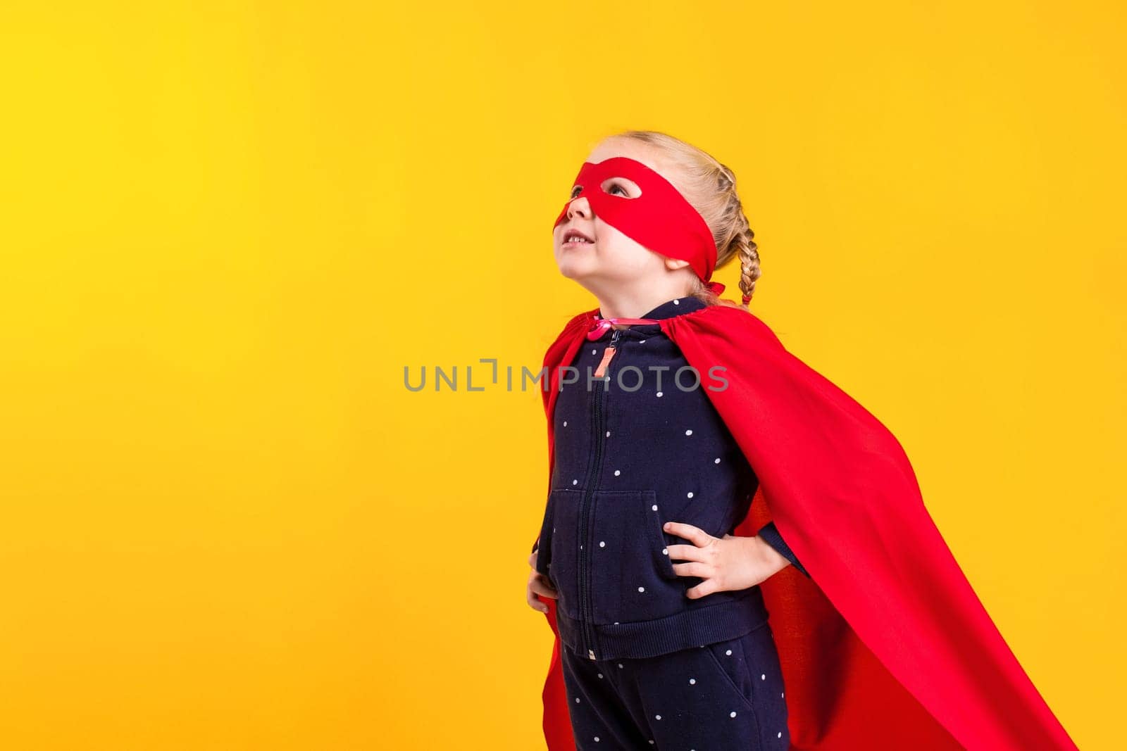 Funny little power superhero child girl in a red raincoat and a mask on a yellow background. Studio photography. Superhero concept.