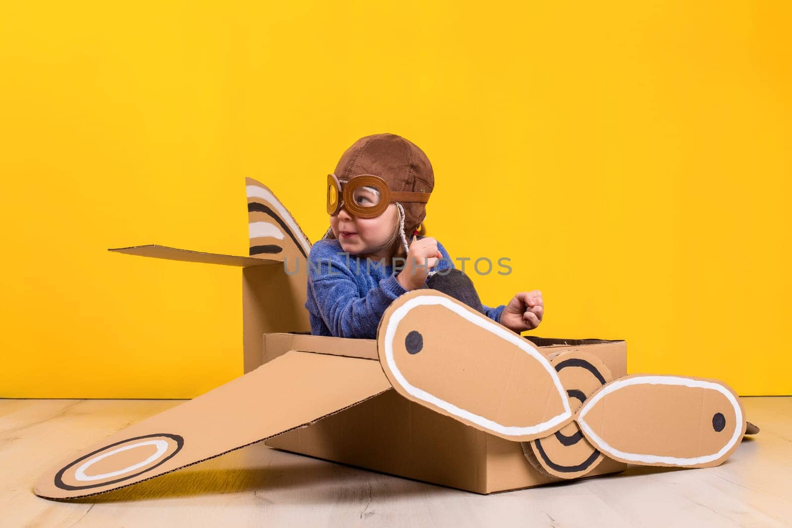 Little dreamer girl playing with a cardboard airplane. Childhood. Fantasy, imagination. Studio photography on a yellow background. Imagination or exploration concept