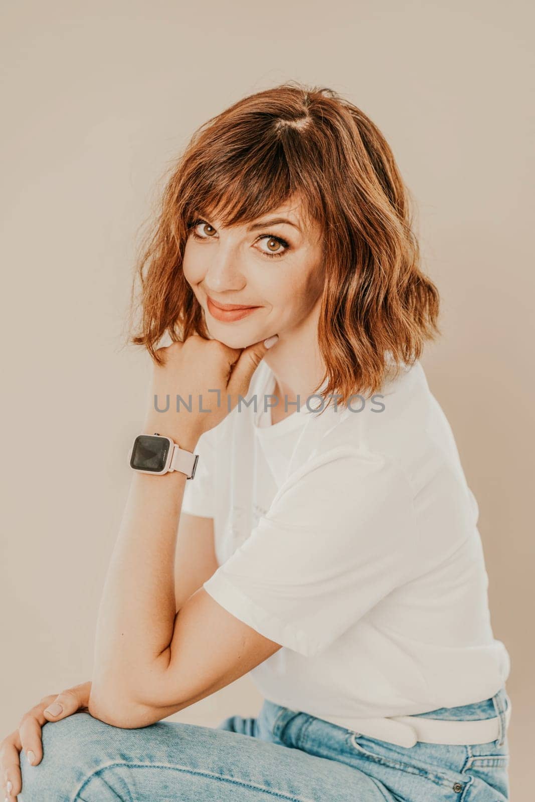 Portrait of brunette white t-shirt. A woman sits on a chair and looks at the camera, business portrait on a beige background, dressed in a white t-shirt and jeans