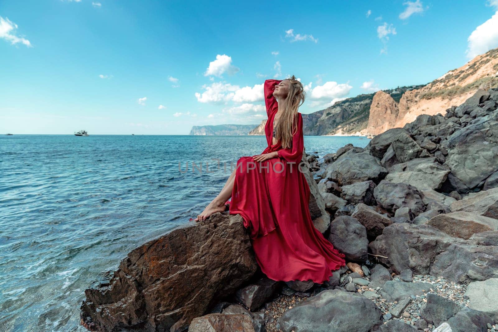 Red dress sea woman. A blonde with flowing hair in a long red dress sits on a rock near the sea. The concept of trips, a photo shoot at the sea by Matiunina